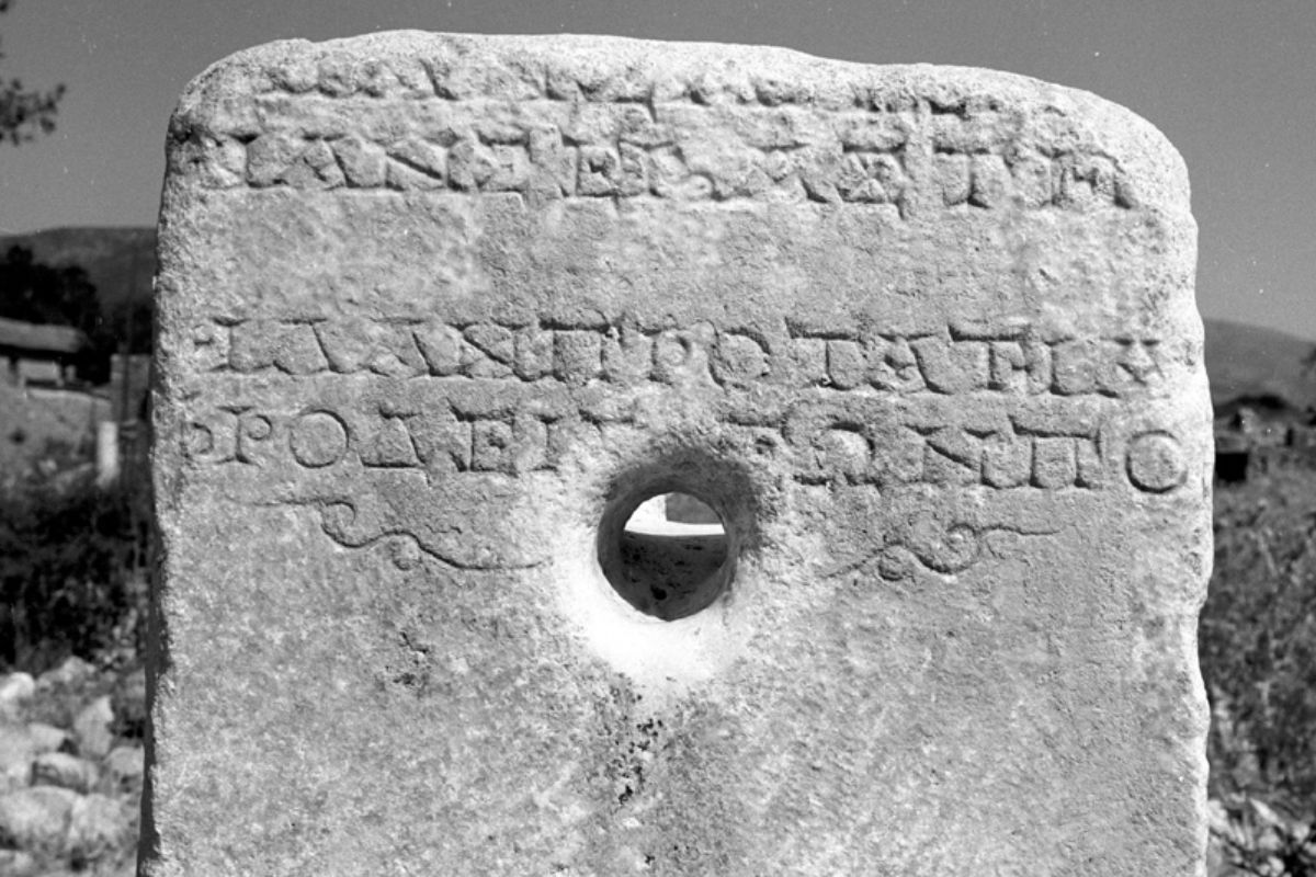 An ancient stone inscription with Greek text, featuring a central circular hole, set against a backdrop of ruins and a mountainous landscape.